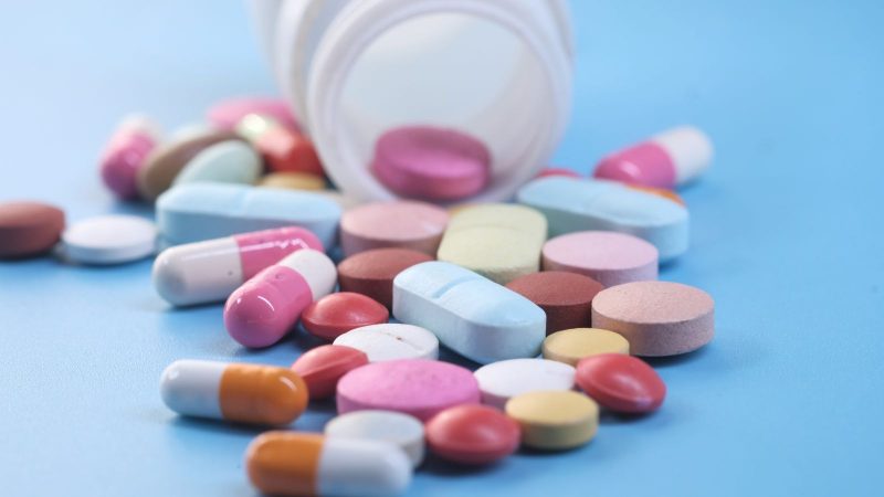 Tadalafila Imagem mostra vários comprimidos de diferentes cores saindo de dentro de uma caixa de remédio sob um fundo azul claro. A imagem serve para ilustrar a matéria que explica o que é medicamento tadalafila.