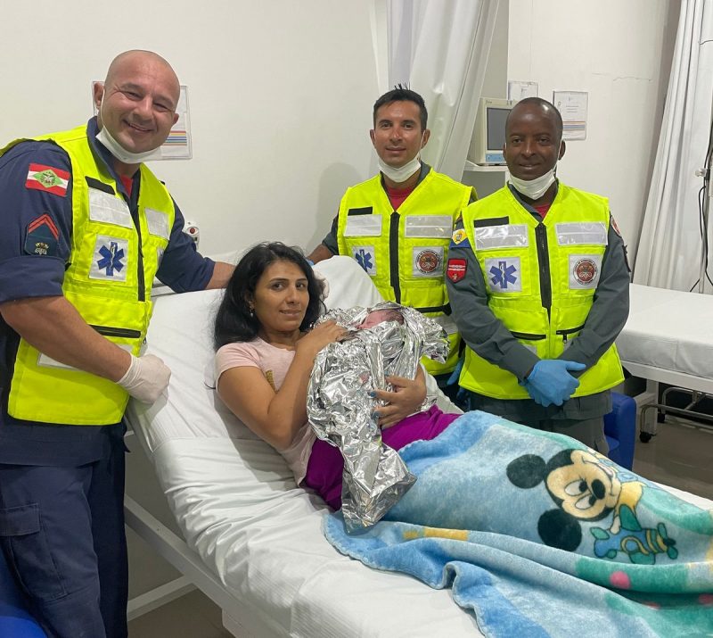 Mãe segura bebê Foto mostra mãe segurando bebê na maca do hospital, com 3 bombeiros em volta