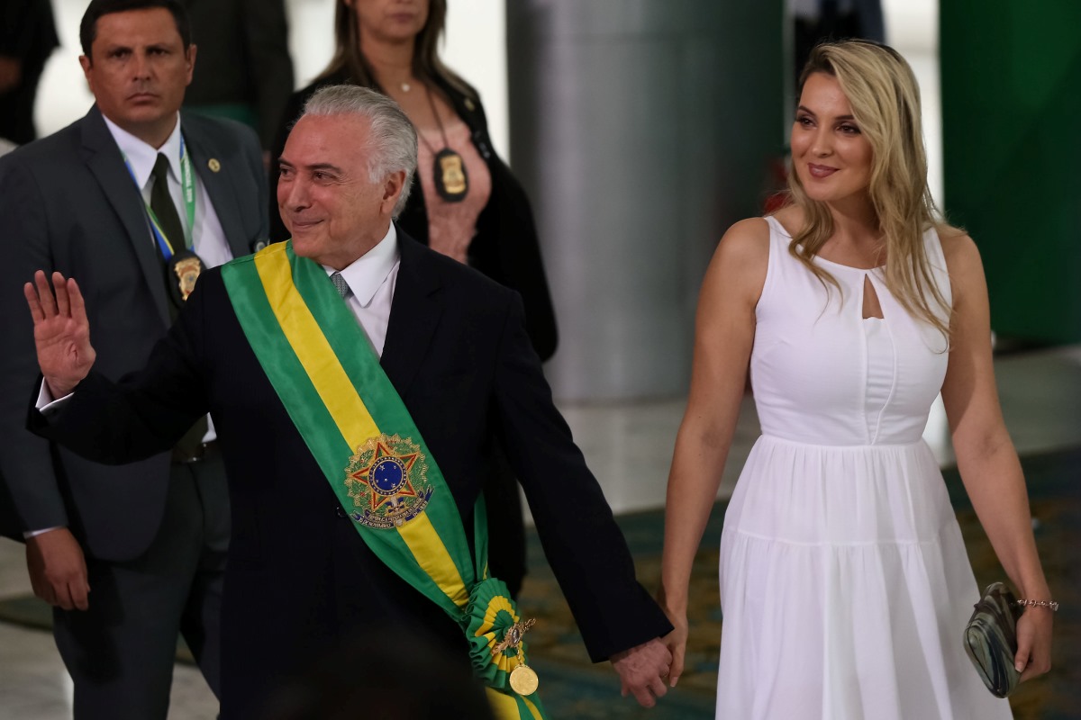 Na imagem, Michel Temer, ex-presidente do Brasil. Ele é um homem branco e grisalho, que sorri e veste terno com uma faixa verde e amarela. Ele está de mãos dadas com Marcela Temer, sua esposa, que veste um vestido branco. Ela é loira, branca e sorri.
