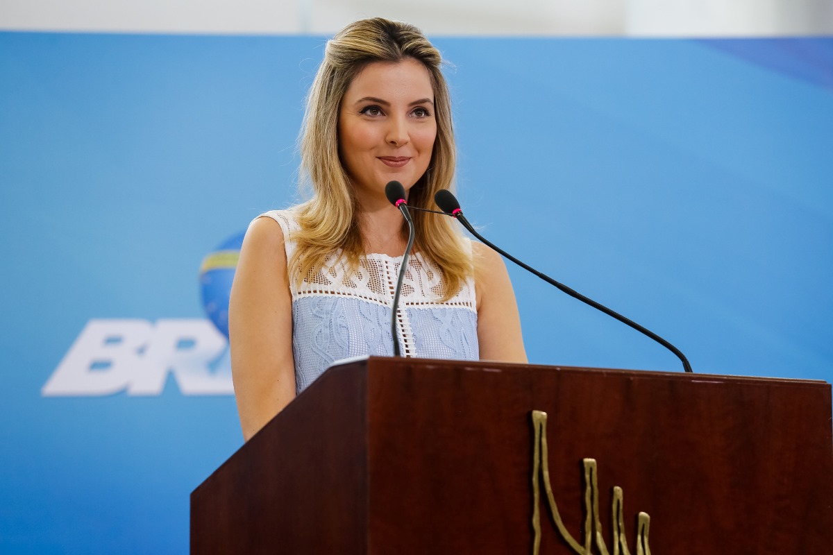 Marcela Temer na Cerimônia de Lançamento do Programa Criança Feliz. Marcela é uma mulher branca, loira, que veste um vestido azul. Ela sorri enquanto discursa em um palanque com microfones. O fundo da imagem é azul e possui a inscrição "Brasil".