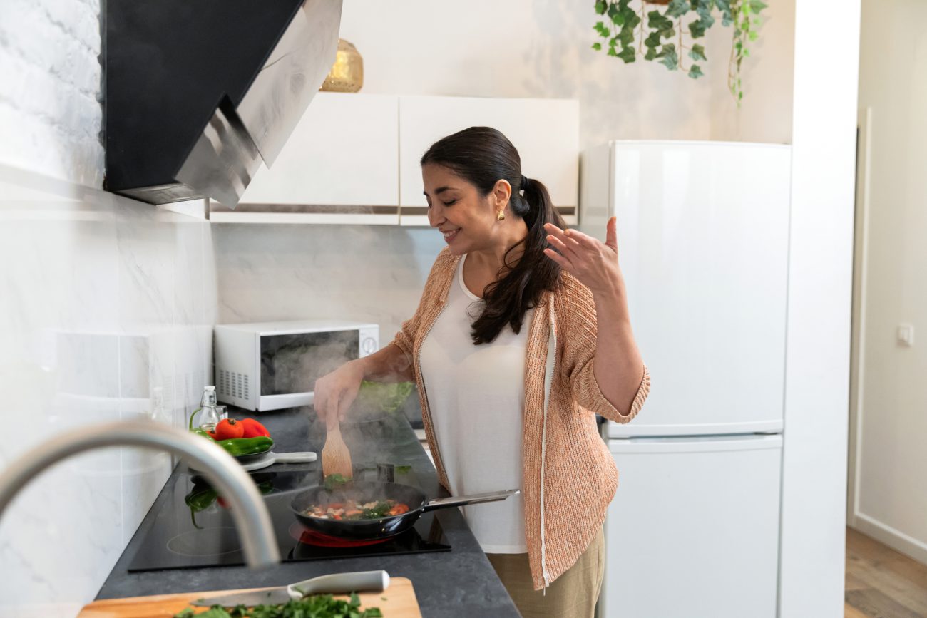 Mulher cozinhando legumes na frigideira em cozinha com exaustor