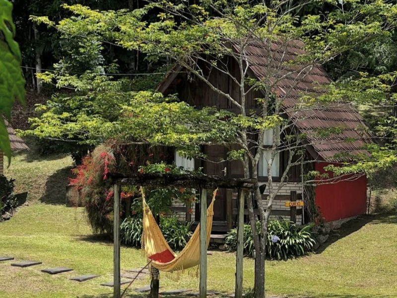 Pousada Rio da Prata em Blumenau Imagem mostra um dos chalés da Pousada Rio da Prata, em Blumenau. Na foto é possível ver parte do chalé, com muitas árvores e natureza ao redor com uma rede na cor amarela em frente