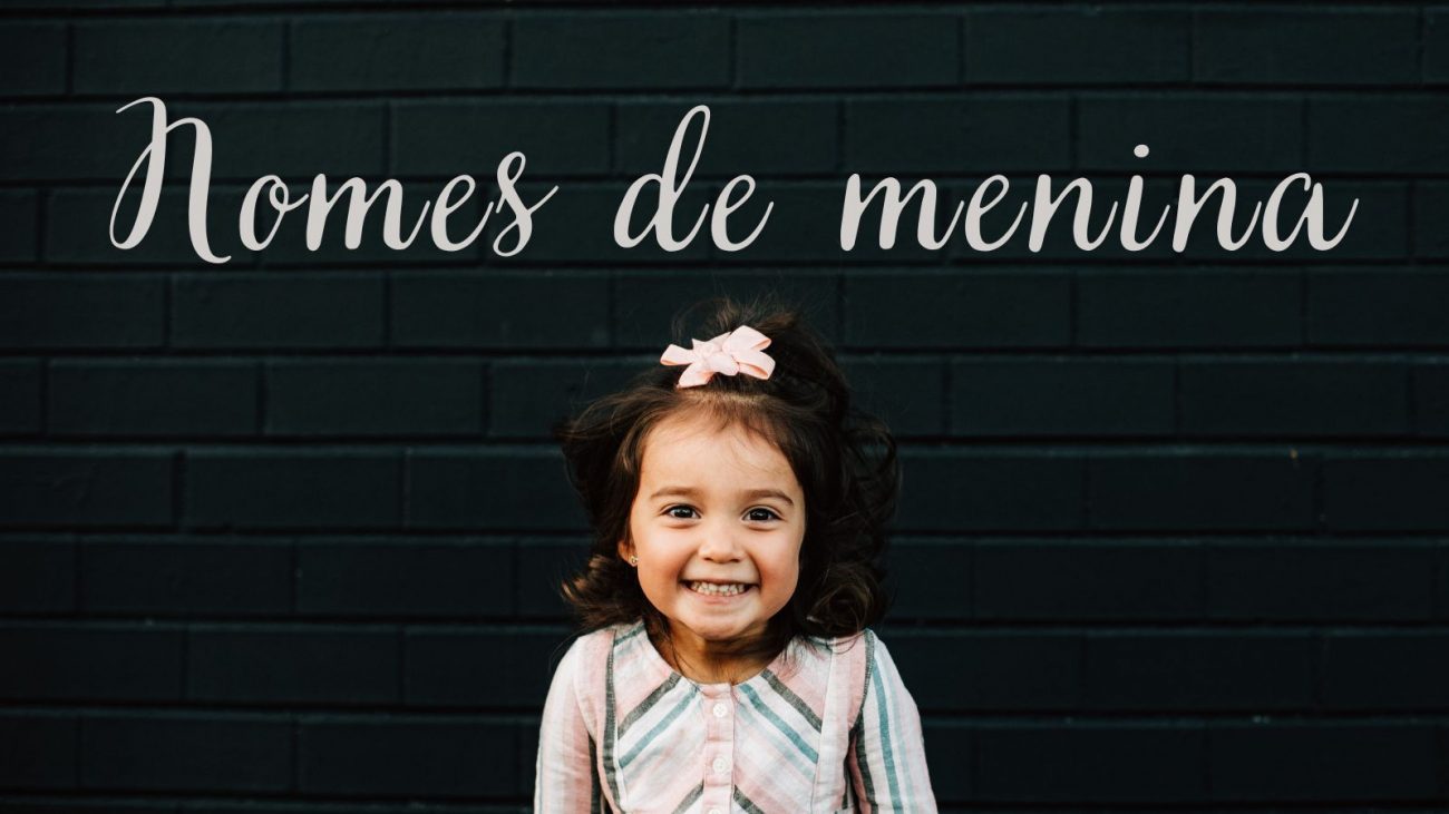 Imagem mostra uma frase escrito "Nomes de menina" e uma menina branca, com o cabelo amarrado em um laço rosa, sorridente, com roupa colorida e cabelos escuros. O fundo é uma parede escura de tijolos. A imagem é utilizada para ilustrar a matéria que traz nomes compostos de menina. 