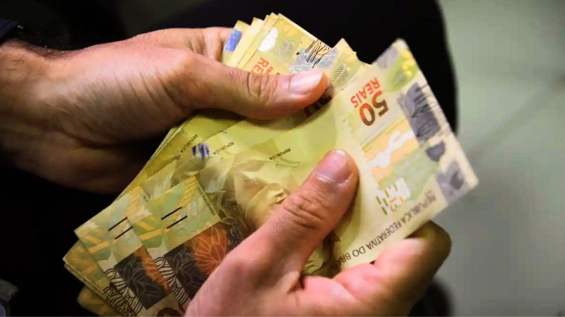 Aprenda a calcular o abono salarial Imagem mostra duas mãos contando várias notas de cinquenta reais para ilustrar o valor pago pelo abono salarial PIS/Pasep