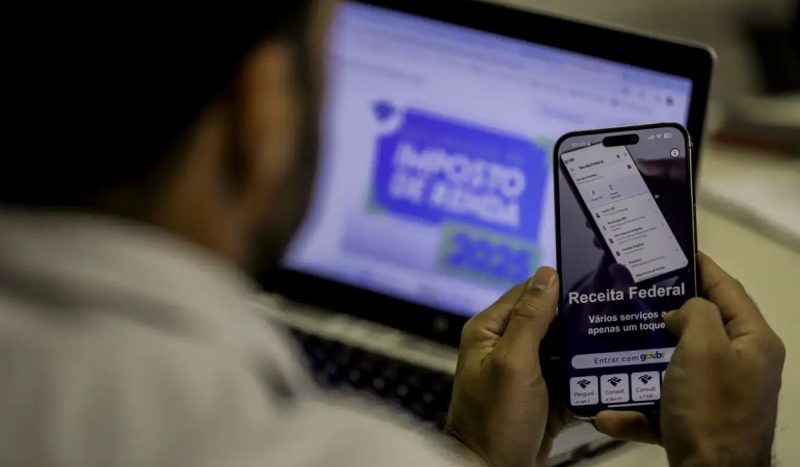 Homem mexendo no aplicativo da Receita Federal pelo celular, com a página do Imposto de Renda aberta no notebook Nova tabela do Imposto de Renda começou a valer no dia 1 de maio