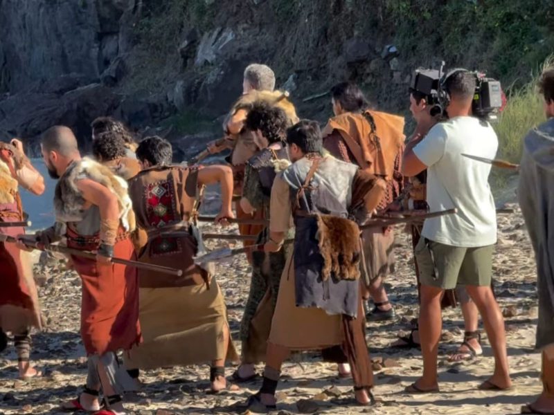 Novela Paulo, o Apóstolo Imagem mostra várias pessoas de costas com roupas de época. Elas estão em uma praia. É possível ver a areia, um pouco do mar e pedras rochosas ao redor
