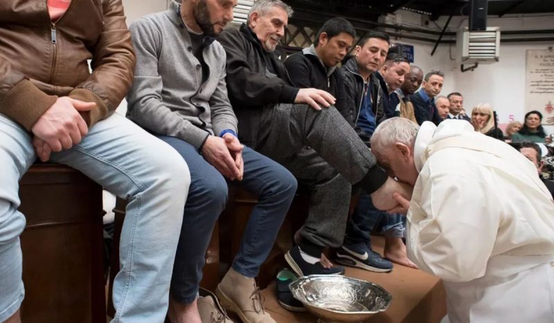 Papa Francisco costumava lavar os pés de detentos, foto mostra Francisco ajoelhava em frente a fileira de homens, com uma bacia de água no chão a sua frente, ele beija o pé de um dos homens