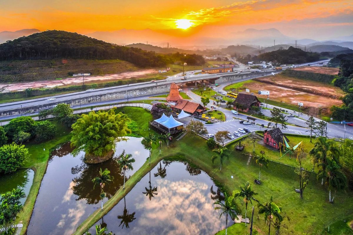 Parque Expoville Joinville Foto aérea do Parque Expoville, em Joinville. Na parte inferior, aparece parte de três lagos, com um extenso gramado ao redor e algumas árvores. Ao fundo, uma rodovia, morros verdes e o pôr do sol. O céu está alaranjado.