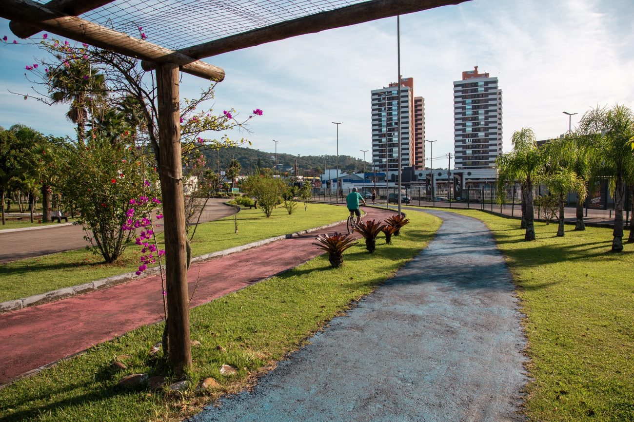 Parque dos Imigrantes Criciúma Foto do Parque dos Imigrantes, em Criciúma. No centro da foto aparece uma pista de caminhada e uma ciclovia, com áreas gramadas ao redor. Há também um pergolado de madeira com flores rosas. Ao fundo, aparecem algumas árvores, prédios e no horizonte morros verdes. O céu está azul.