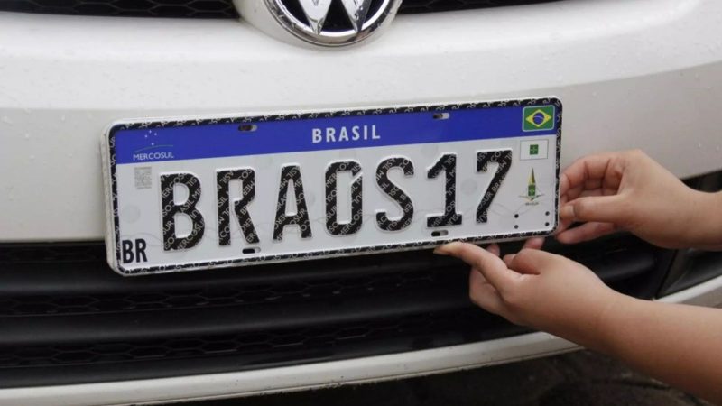 na foto aparece uma placa do carro sendo colocada por uma pessoa 