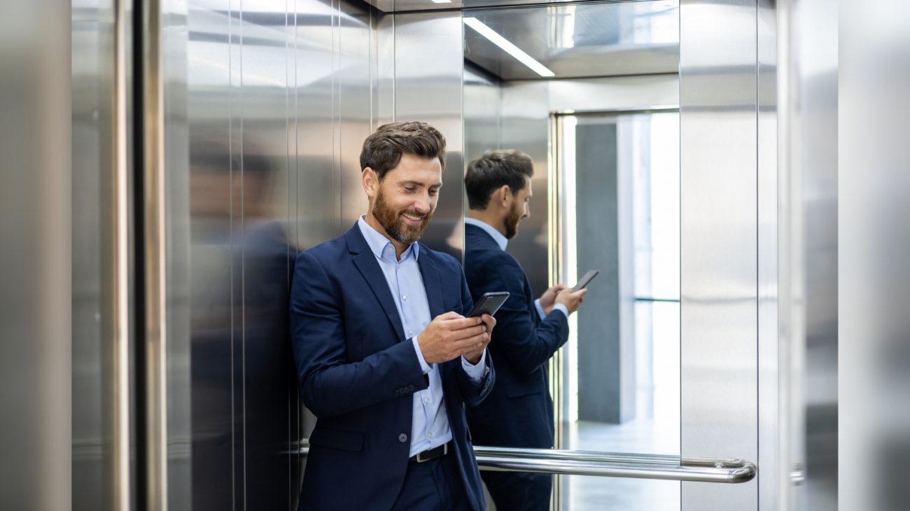 Pessoa com celular na mão Pessoa de terno azul e blusa azul bebê com celular na mão sorrindo dentro de elevador por que os elevadores têm espelhos