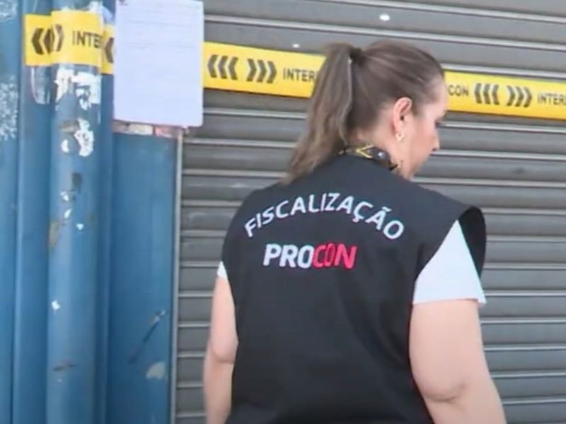 Loja de pneus de Blumenau Imagem mostra uma mulher de costas que faz parte da fiscalização do Procon de Blumenau. Ela usa um colete preto com a escrita FISCALIZAÇÃO PROCON. Ao fundo dela tem uma porta da loja de pneus de Blumenau fechada