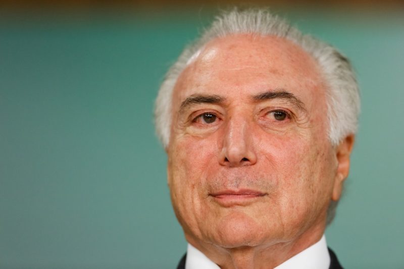 Imagem de Michel Temer, ex-presidente do Brasil. Michel Temer é um homem branco, grisalho e idoso, de olhos castanhos. A fotografia foca em seu rosto e o fundo da imagem é em tons de verde acinzentado.
