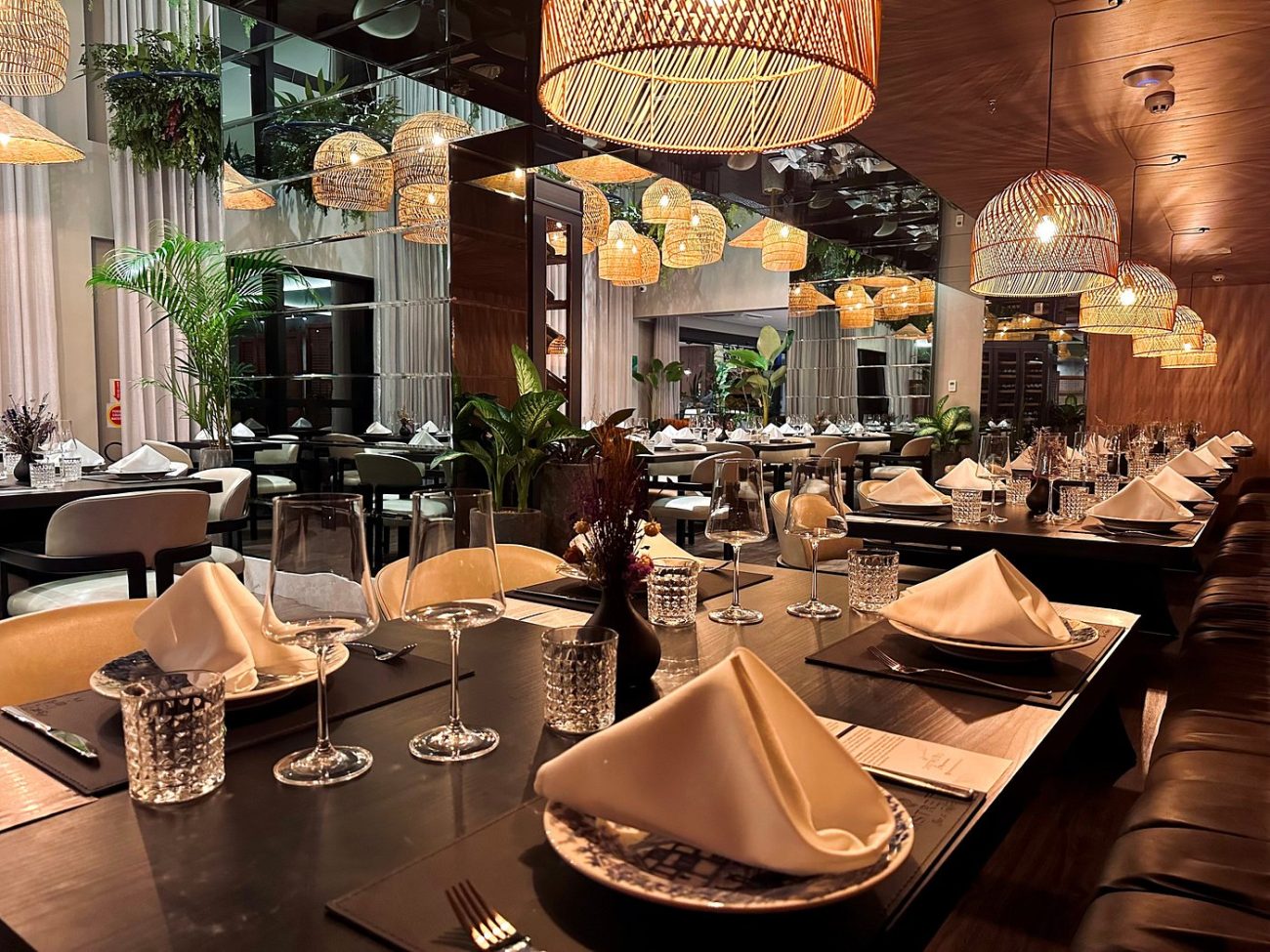 Foto interna do restaurante com mesas de madeira escura, sobre as quais estão pratos e guardanapos de pano e taças de vidro. Uma das paredes tem um grande espelho e há vários lustres de sisal.