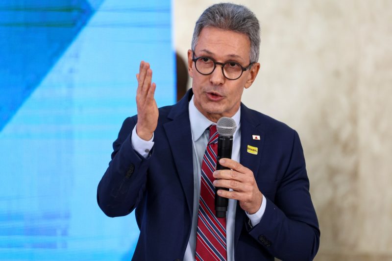 Imagem de Romeu Zema, que ilustra matéria no Portal NDMais sobre o político. Zema é um homem branco, grisalho, que veste terno, gravata vermelha listrada e utiliza óculos preto. Segura um microfone nas mãos.