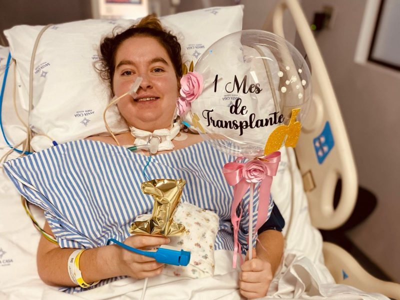 História de Isis, transplante de pulmão Imagem mostra uma jovem deitada em uma cama de hospital segurando um balão. Ela tem uma sonda no nariz além de uma traqueo no pescoço. Ela usa uma roupa listrada e foto aconteceu após transplante de pulmão