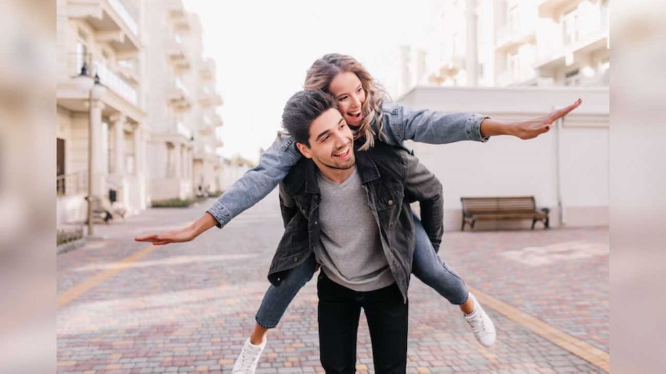 Homem com jaqueta preta carregando mulher que está de braços abertos e jaqueta jeans nas costas// signos que combinam