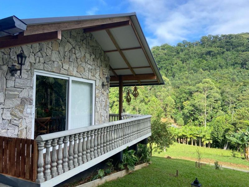 Cabana de Pedra Imagem mostra uma cabana de pedras, em cima de um morrinho. Ela tem janelas brancas e parapeito na cor branca também. O céu está bem azul e há muito verde e muitas árvores na paisagem