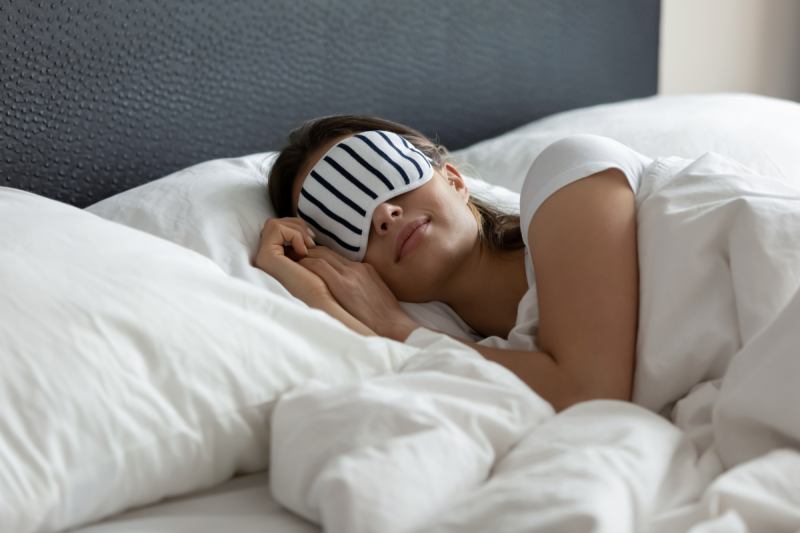 Turismo do Sono SC Mulher jovem, de pele clara e cabelos castanhos cumpridos deitada em uma cama com travesseiros e edredom brancos. Ela usa uma máscara de dormir branca com listras pretas.