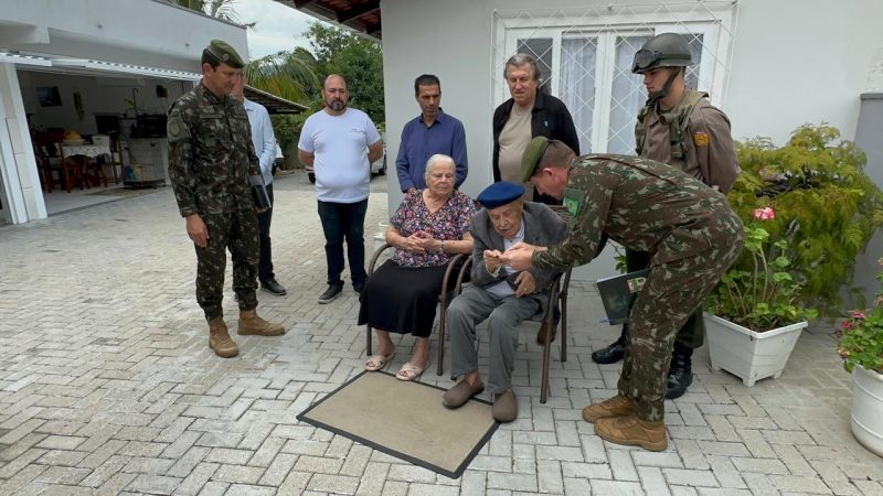 Recebimento da homenagem Foto mostra sargento Hugo, único veterano da Segunda Guerra Mundial vivo em SC, sentado enquanto recebe homenagem de militar.