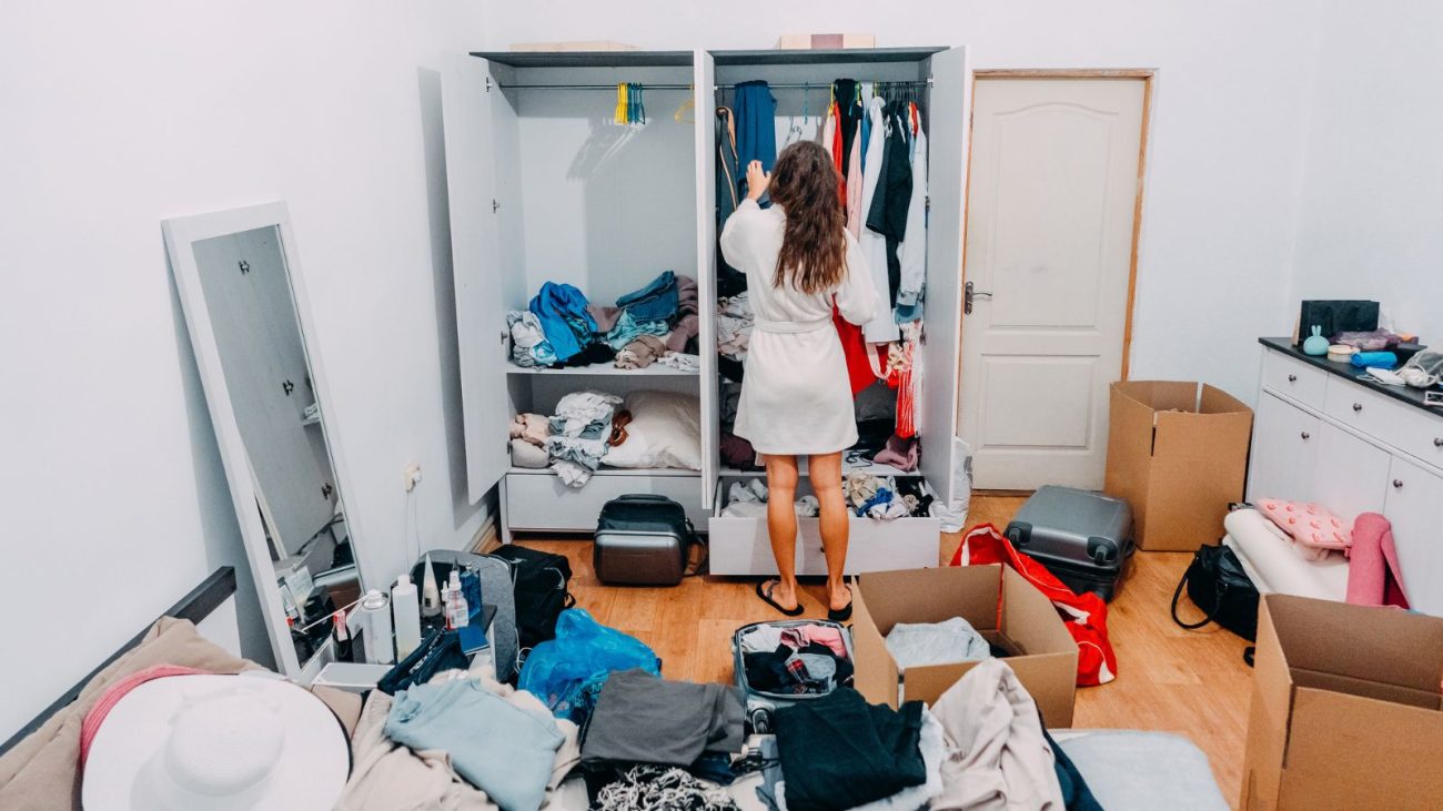 Mulher organizando roupas no guarda-roupa em um quarto bagunçado, com várias roupas e caixas espalhadas — situação comum entre pessoas desorganizadas.