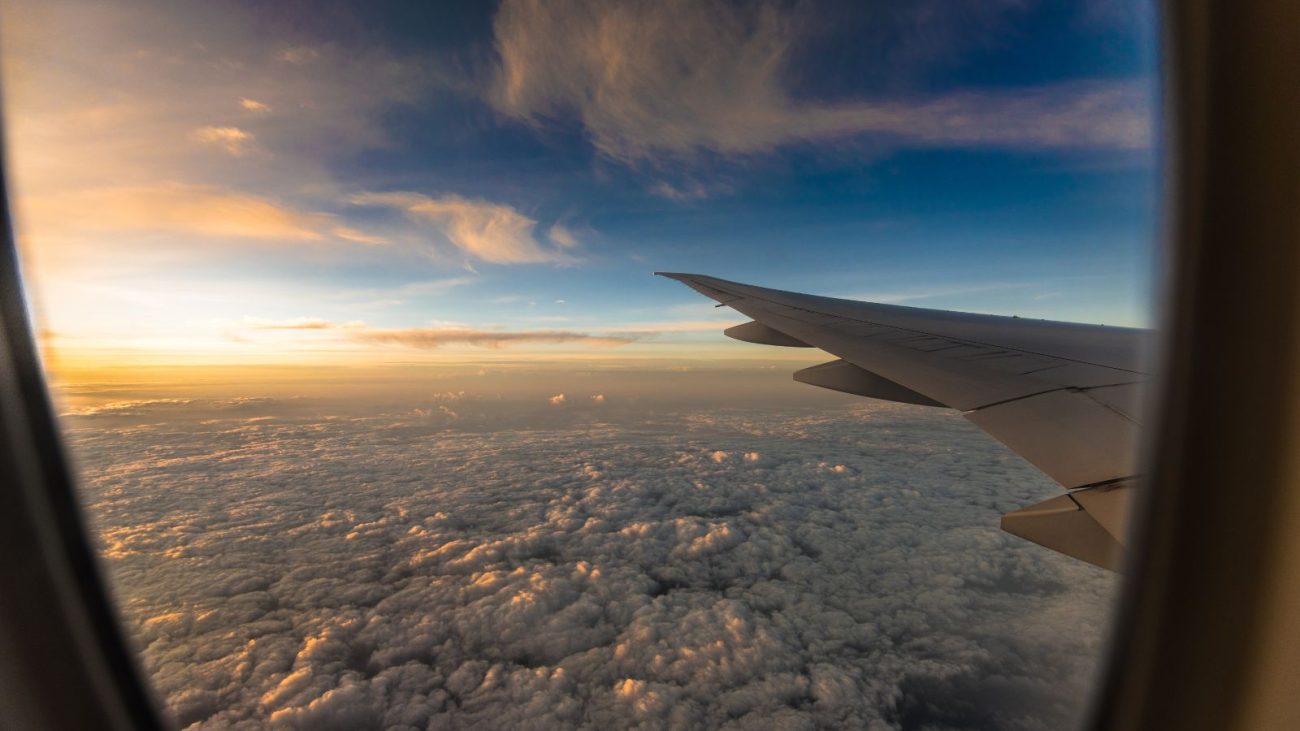 Vista da janela de avião durante o voo com nuvens e céu ao entardecer, transmitindo sensação de viagem e aventura aérea.