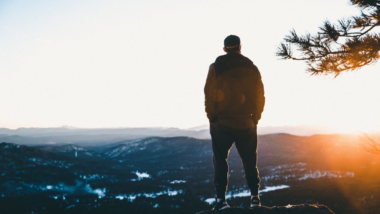 Homem parado em cima de um morro observando o pôr do sol no horizonte