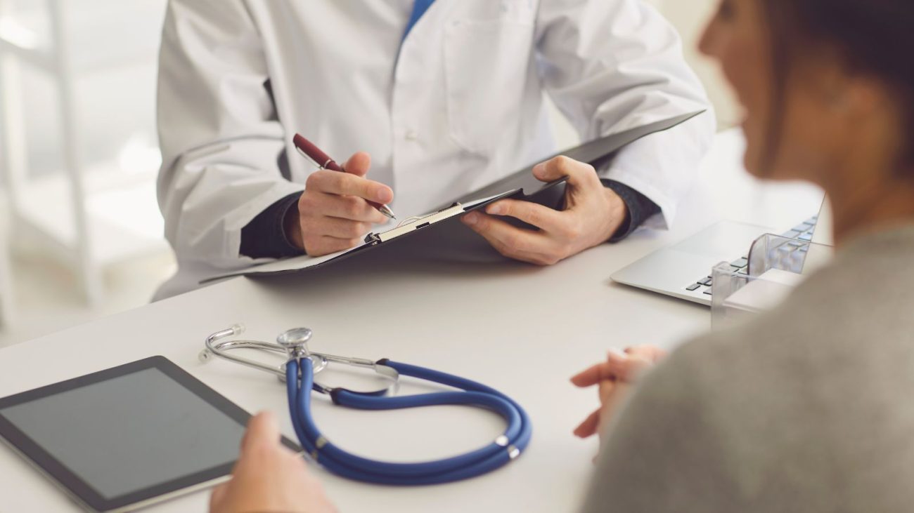 Médico com jaleco branco conversa com uma paciente em consultório, enquanto faz anotações em uma prancheta. Sobre a mesa, há um estetoscópio, um tablet e um notebook. A imagem representa uma consulta médica, onde são discutidos os exames obrigatórios em um check-up de rotina.