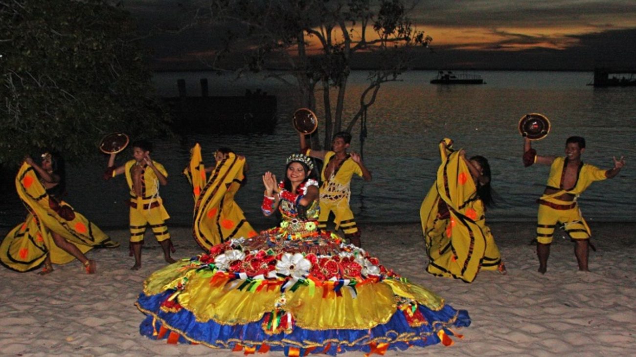 Dança tradicional indígena na praia ao pôr do sol, com mulheres e homens usando roupas coloridas e chapéus típicos, celebrando a cultura local.