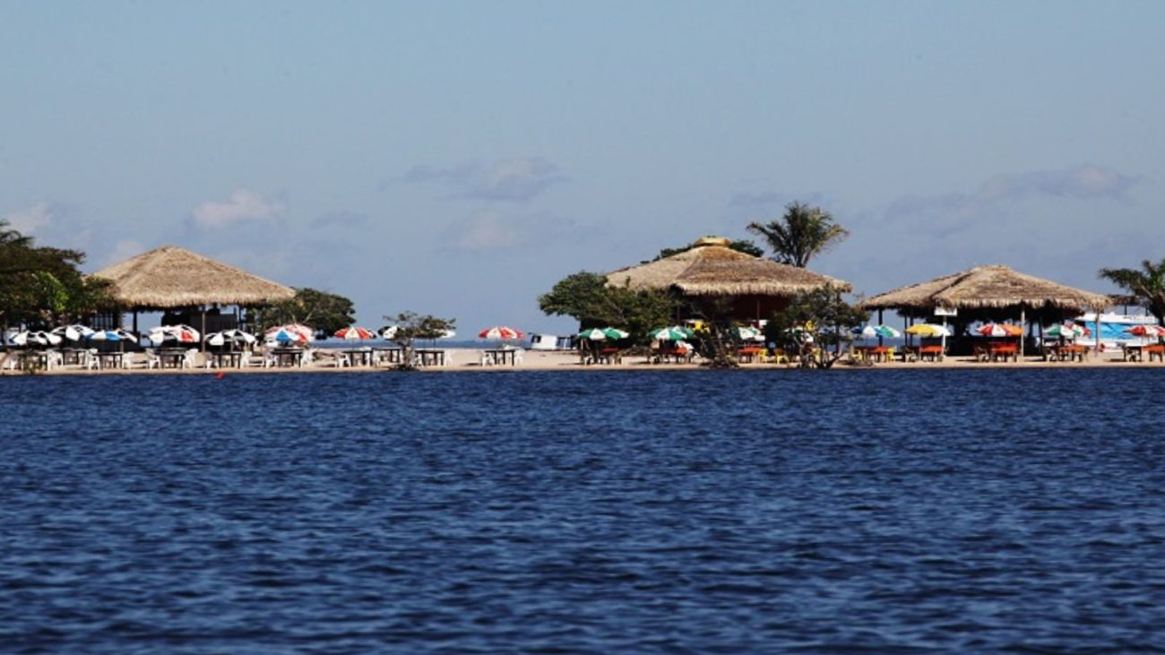 Imagem de uma praia com restaurantes de palha e cadeiras de praia sob guarda-sóis coloridos, vista para o mar azul claro sob céu parcialmente nublado.