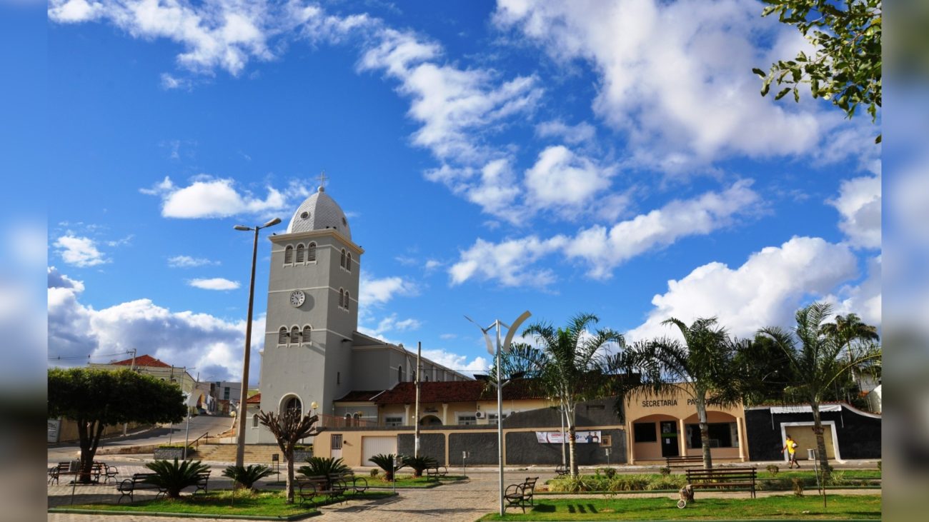 Imagem da cidade de Araçuaí, conhecida por ser uma das mais quentes do Brasil, com céu azul, nuvens brancas, árvores, praça e edifícios históricos.