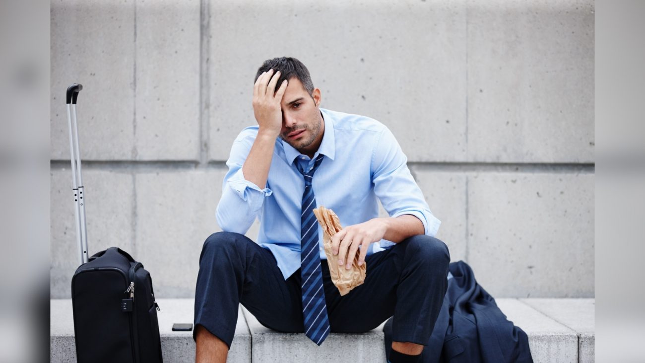 Homem sentado em calçada com mala e bolsa ao lado segurando pacote