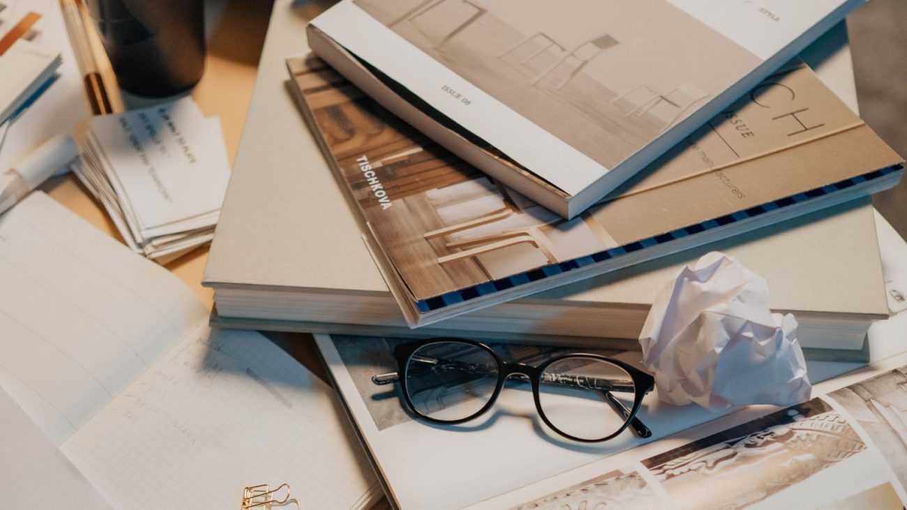 Mesa de trabalho com livros, óculos, papéis amassados e anotações, simbolizando estudo ou pesquisa acadêmica ou profissional.