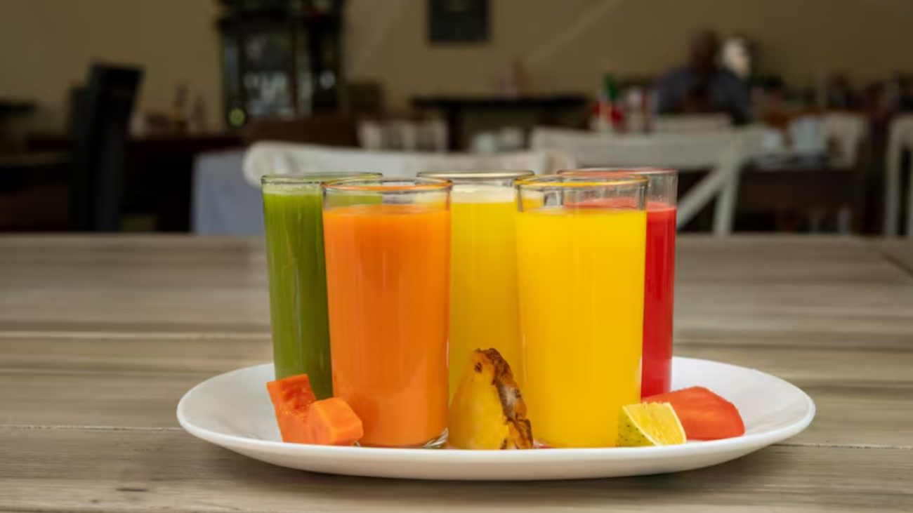 Vários copos de suco colorido com frutas ao lado em uma bandeja branca: verde, laranja, amarelo e vermelho, em um ambiente de restaurante.
