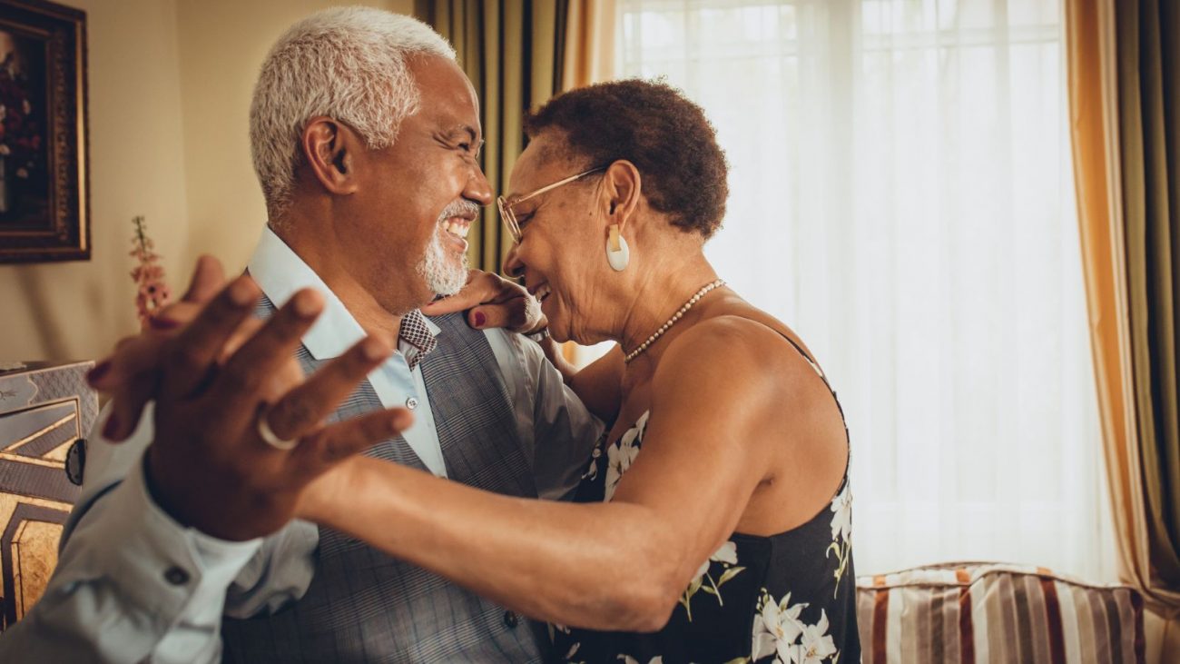 Imagem mostra um casal mais velho, dançando abraçado na sala de casa, sorrindo, para representar as bodas de jequitibá. O homem está com uma roupa social clara e a mulher com um vestido preto florido, utilizando óculos de grau, brincos e colar. Ele aparece apenas com a aliança como acessório.