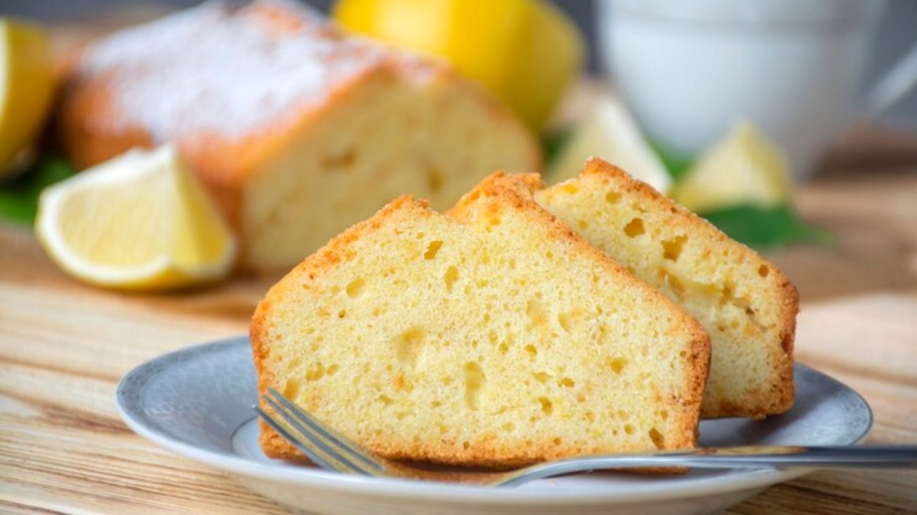 Prato com duas fatias de bolo de limão dourado e macio, servido com um garfo. Ao fundo, o restante do bolo inteiro com cobertura polvilhada e limões cortados decorando a cena sobre uma superfície de madeira.