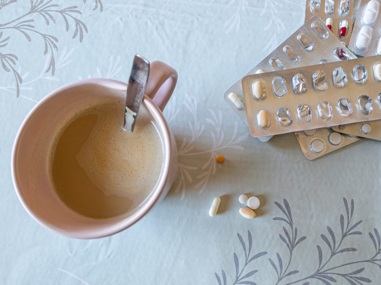 Xícara de café com cartelas de remédio e comprimidos espalhados sobre a mesa para alertar para os riscos da interação entre cafeína e medicamentos