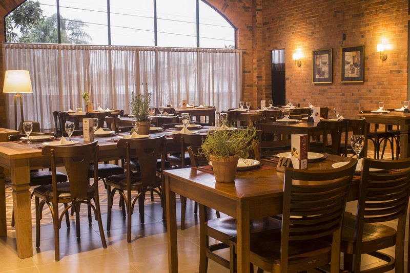 Ambiente interno da Cantina Vettorazzi, em Criciúma. A imagem mostra um salão amplo com mesas e cadeiras de madeira, abajur, parede de tijolos à vista e uma grande janela de vidro com a parte superior em formato de semicírculo. 