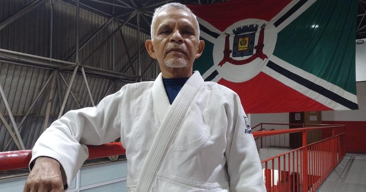 Imagem de um home de 64 anos, em frente abandeira de Criciúma, para fazer referência ao fato de que ele representa município em várias competições 