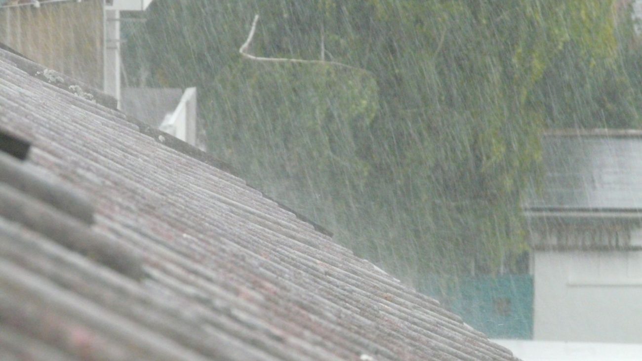 Chuva forte caindo sobre telhados de casas em uma área urbana, com árvores ao fundo desfocadas pela intensidade da água. A imagem representa os efeitos de uma frente fria que provoca temporais e mudanças bruscas no clima.