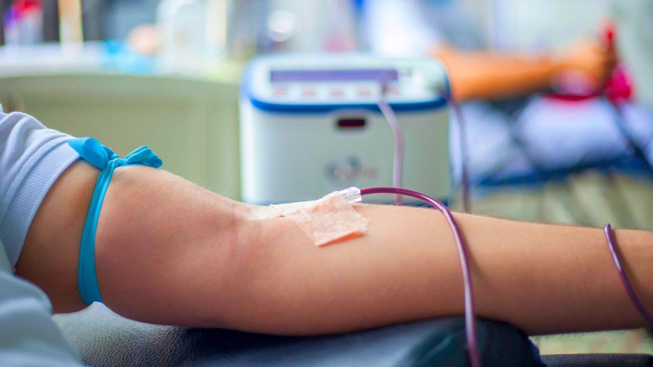Imagem mostra um braço esticado fazendo doação de sangue, com uma fita azul amarrada acima do antebraço, com uma agulhada enfiada na veia na parte contrário ao cotovelo, e um tubo transparente retirando o sangue vermelho de dentro do braço.
