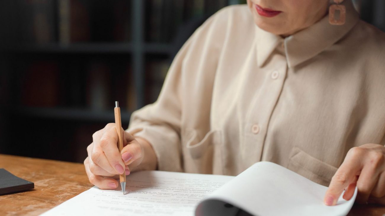 Imagem mostra uma mulher branca, com batom rosa, camisa marrom-claro, escrevendo em um papel com uma lapiseira amarela para ilustrar o concurso público que abriu no TCU. 