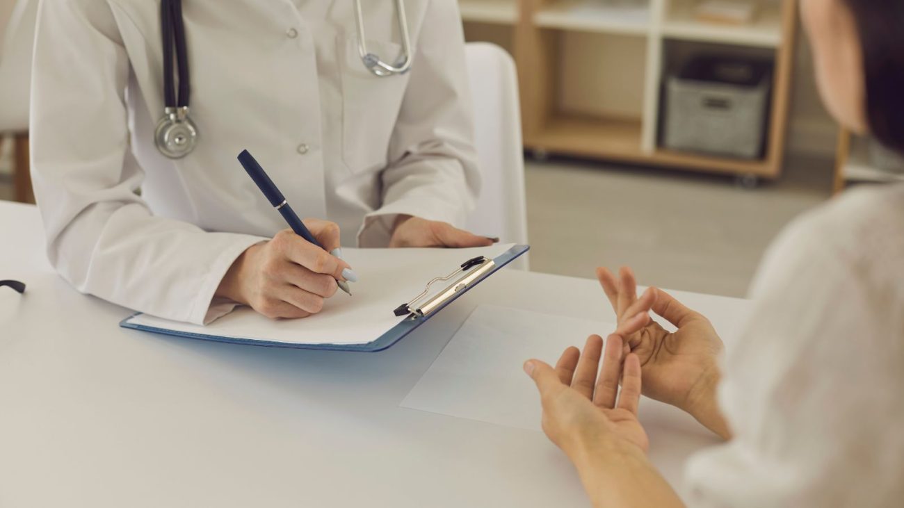 Médico com jaleco branco conversa com uma paciente em consultório, enquanto faz anotações em uma prancheta. A imagem representa uma consulta médica, onde são discutidos os exames obrigatórios em um check-up de rotina.