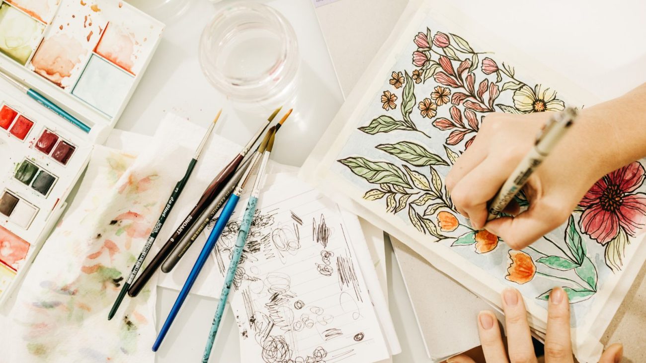 Pessoa criando uma arte com aquarela e tinta, com pincéis, papel e tintas ao lado, em um ambiente criativo e artístico.