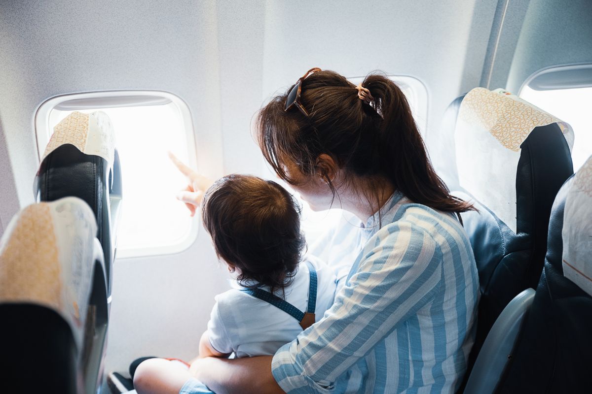 Foto ilustrativa da matéria "Documentos para viajar de avião". Na foto, uma mulher jovem, de cabelos castanhos, longos e presos está sentada em uma poltrona de avião com um menino pequeno no colo. Ambos estão olhando pela janela e ela aponta algo com o dedo.