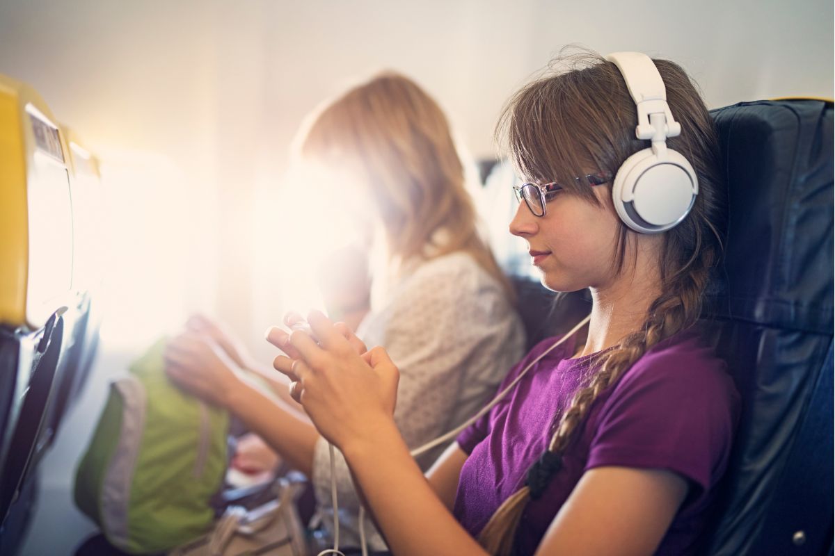 Foto de uma adolescente de cabelos loiros, compridos e com uma trança. Ela está sentada na poltrona de um avião, tem um fone de ouvido e mexe do celular. Ao lado dela, uma mulher loira desfocada.