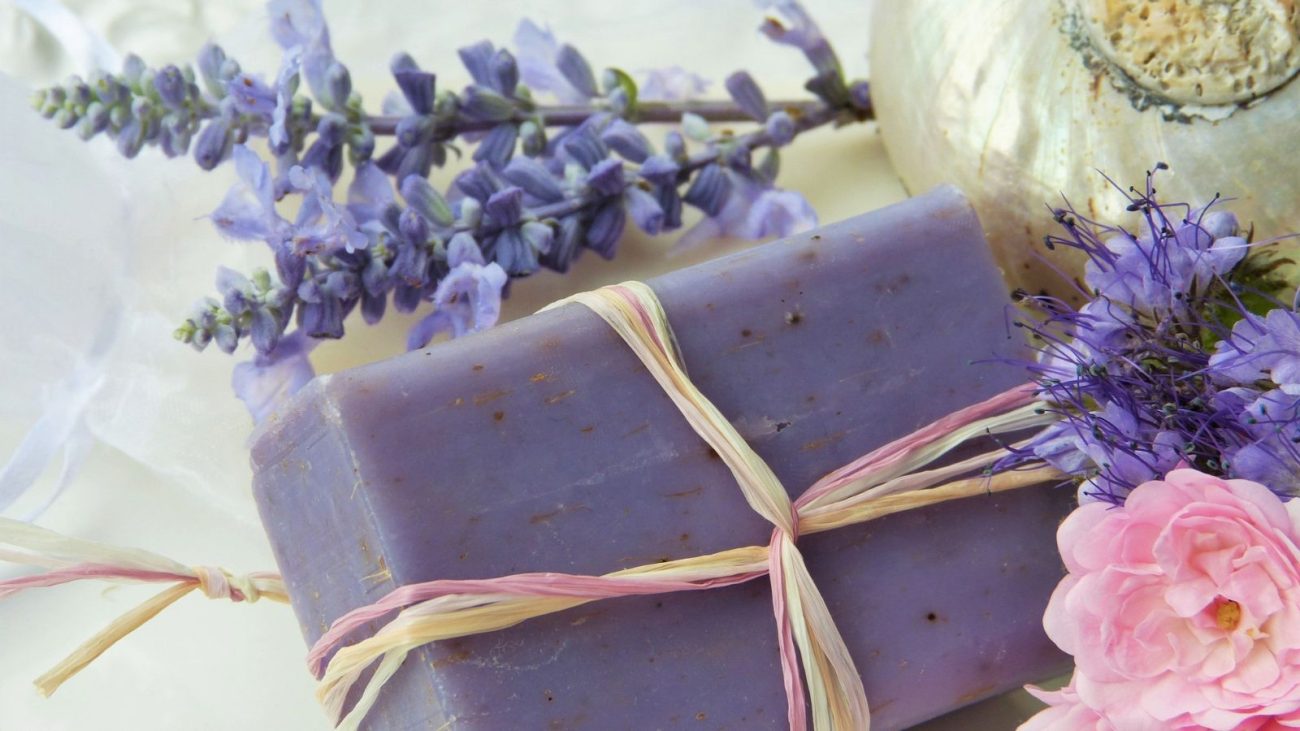 Sabão artesanal de lavanda com flores de lavanda e rosas ao redor, ideal para cuidados com a pele e aromaterapia, em ambiente relaxante.