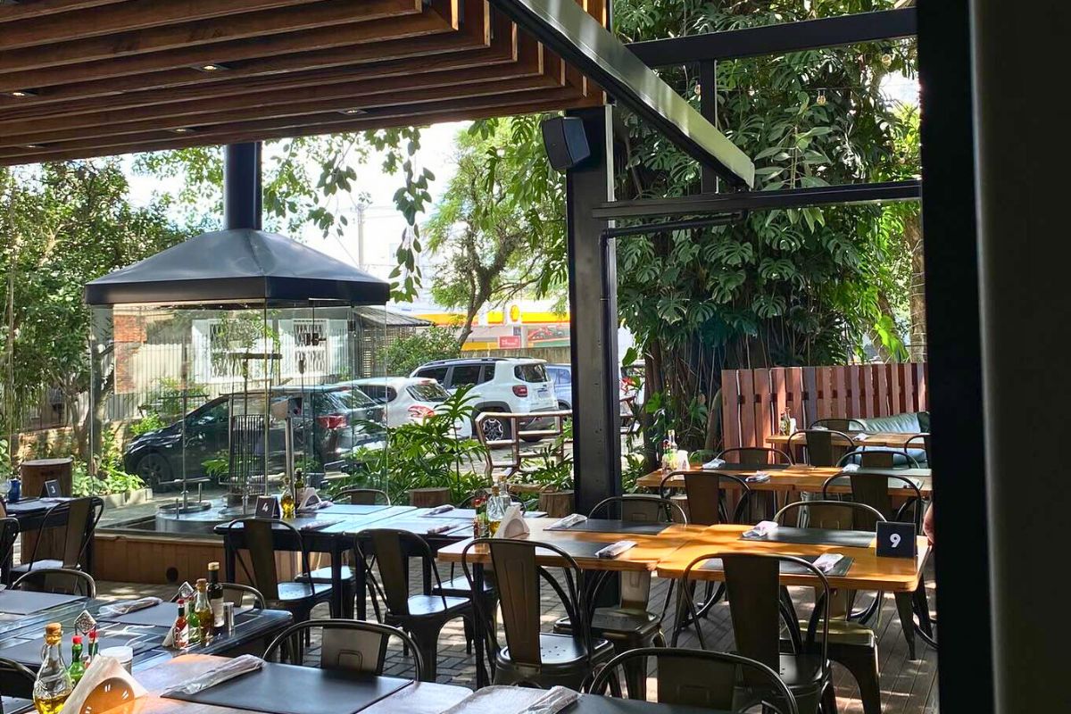 Foto do ambiente externo do El Tarquin. A imagem mostra várias mesas de madeira com cadeiras pretas e uma estrutura preta metálica ao fundo. Aparecem também árvores e vegetações ao redor do espaço. 