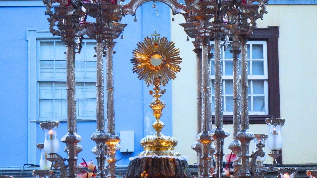 Imagem mostra um ostensório dourado com a hóstia consagrada exposta no centro, sustentado por uma estrutura ornamentada de prata durante a procissão de Corpus Christi. Ao fundo, vê-se a fachada de prédios coloniais em tons de azul e amarelo, com janelas fechadas. A imagem representa a adoração ao Santíssimo Sacramento nas ruas, um dos momentos mais solenes da celebração católica.