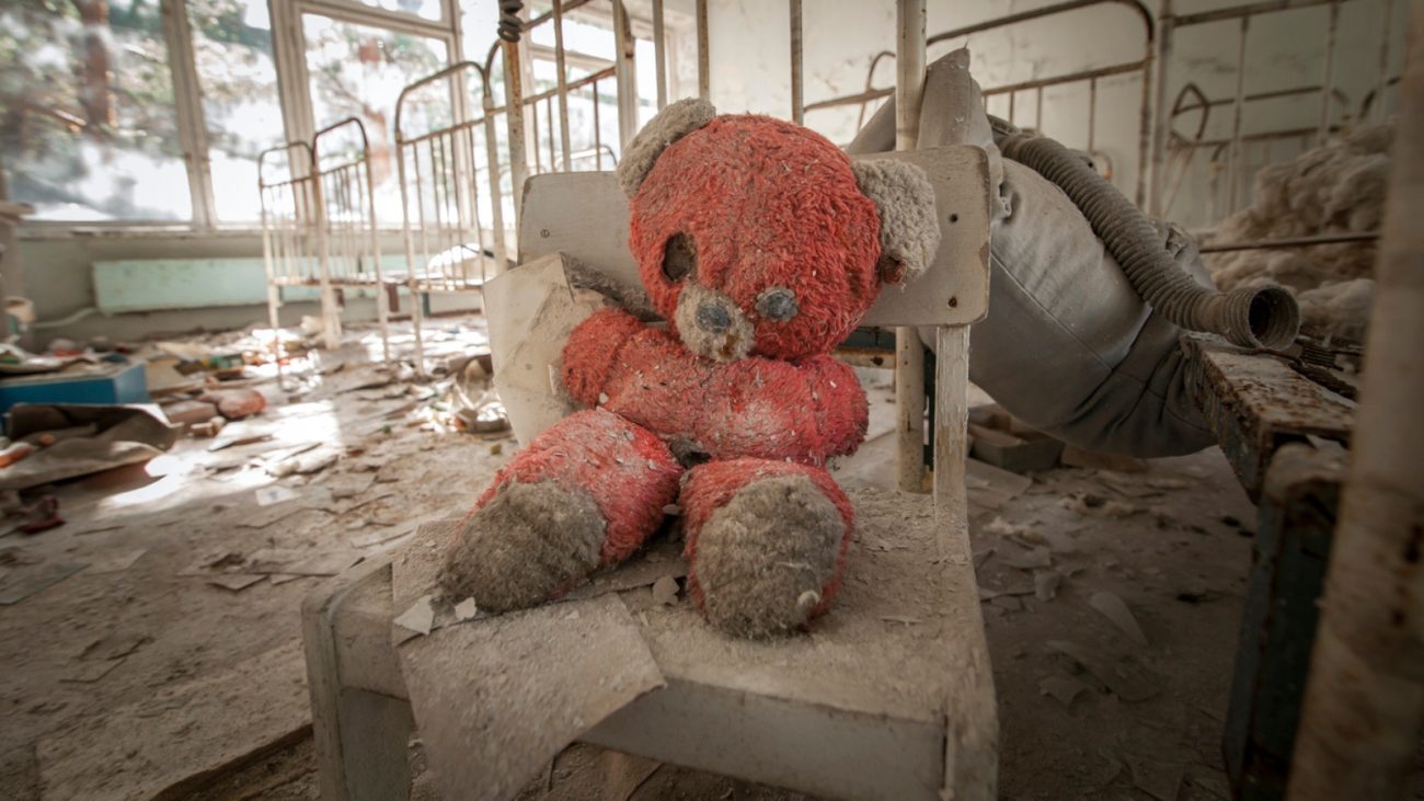 Ursinho de pelúcia vermelho, sujo e rasgado, abandonado em uma cadeira de madeira em um quarto infantil destruído, com berços enferrujados e entulhos ao fundo. A cena sugere um local afetado por catástrofe ou abandono